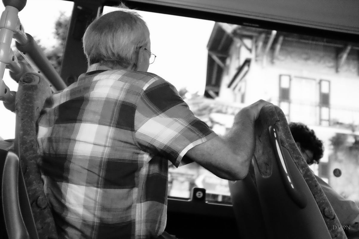 Un homme âgé en chemise à carreaux, vu de dos, tenant un siège dans un bus.