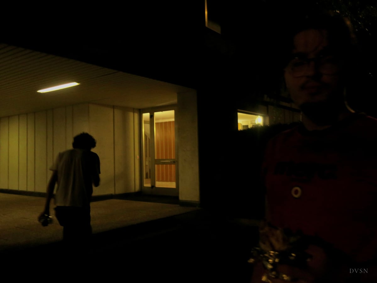 Une silhouette marche vers une entrée éclairée la nuit, tandis qu'un visage à demi éclairé observe depuis le bord droit.