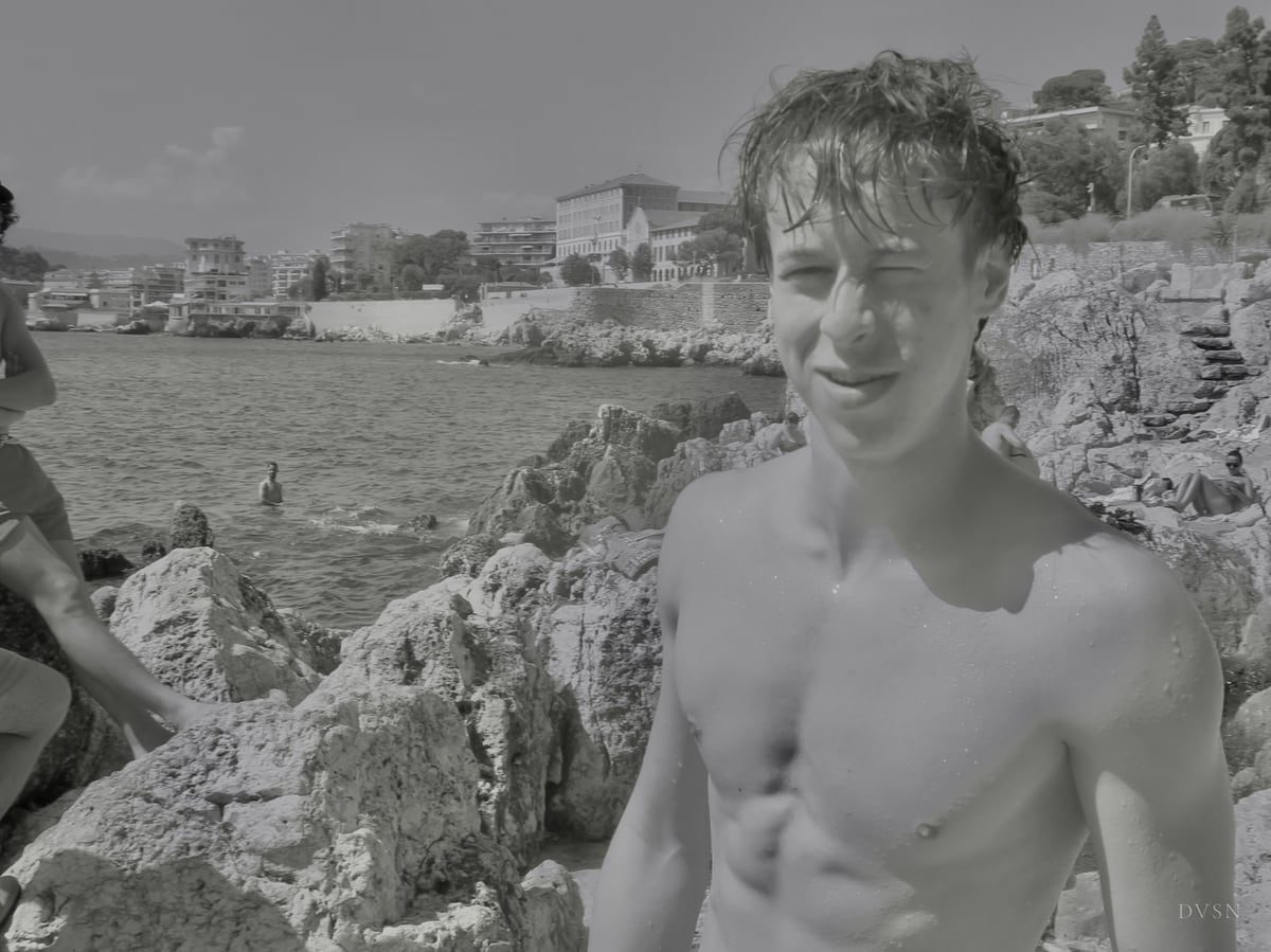 Un jeune homme torse nu, cheveux mouillés, souriant sur des rochers au bord de la mer.