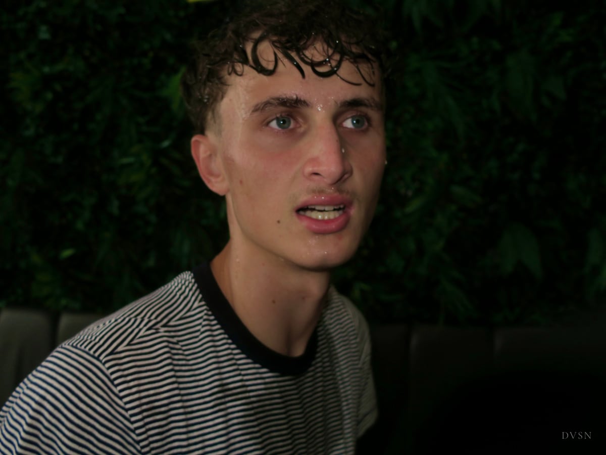 Portrait serré d'un jeune homme aux boucles mouillées et t-shirt rayé, éclairé au flash devant un feuillage sombre.