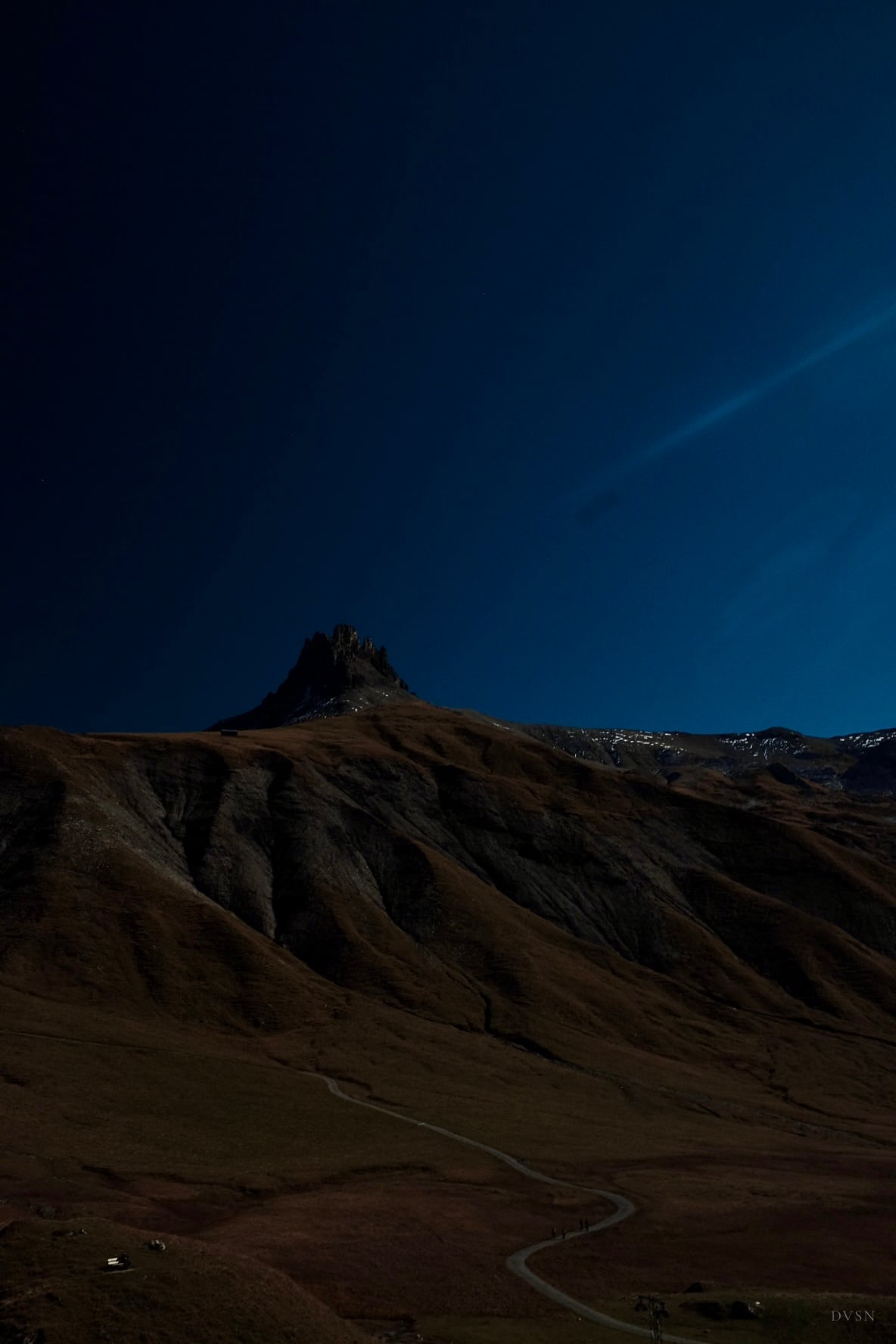 Un pic rocheux sur un ciel bleu profond au-dessus de pentes sombres, un chemin sinueux et de minuscules marcheurs en contrebas.