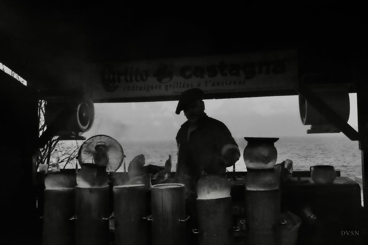 Un vendeur de châtaignes en silhouette derrière son stand, la vapeur qui monte, l'eau et le ciel pâles au loin.