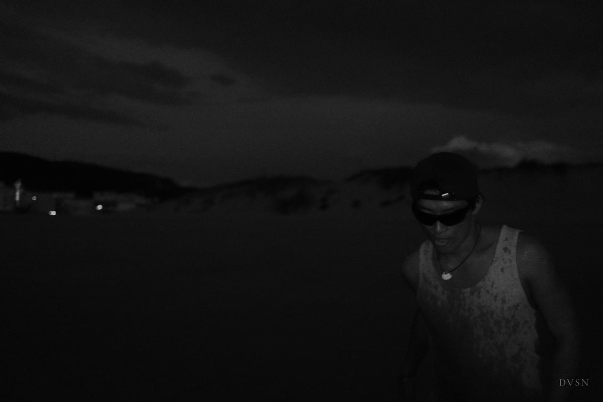 Un homme en casquette et lunettes de soleil émerge d'une obscurité presque totale au bord de l'eau, la nuit.