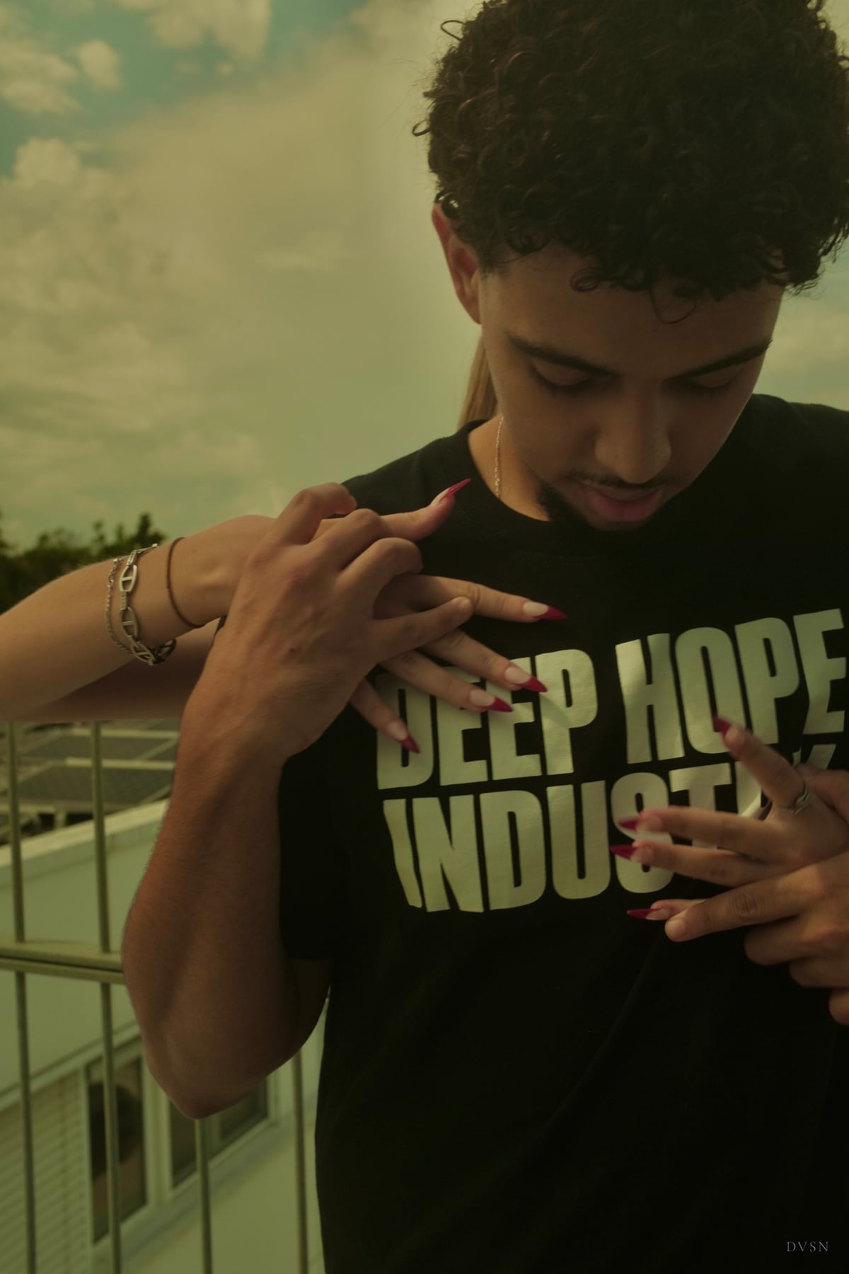 Un jeune homme baisse les yeux tandis que des mains aux longs ongles rouges traversent son t-shirt noir imprimé depuis derrière lui.