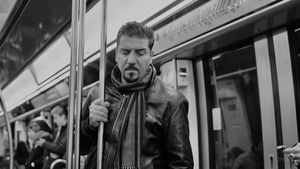 Un homme en blouson de cuir et écharpe tient une barre dans une rame de métro, les yeux baissés.