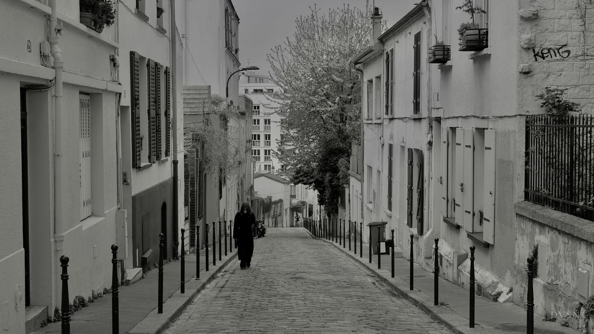 Une silhouette en long manteau sombre descend une rue pavée bordée de potelets.