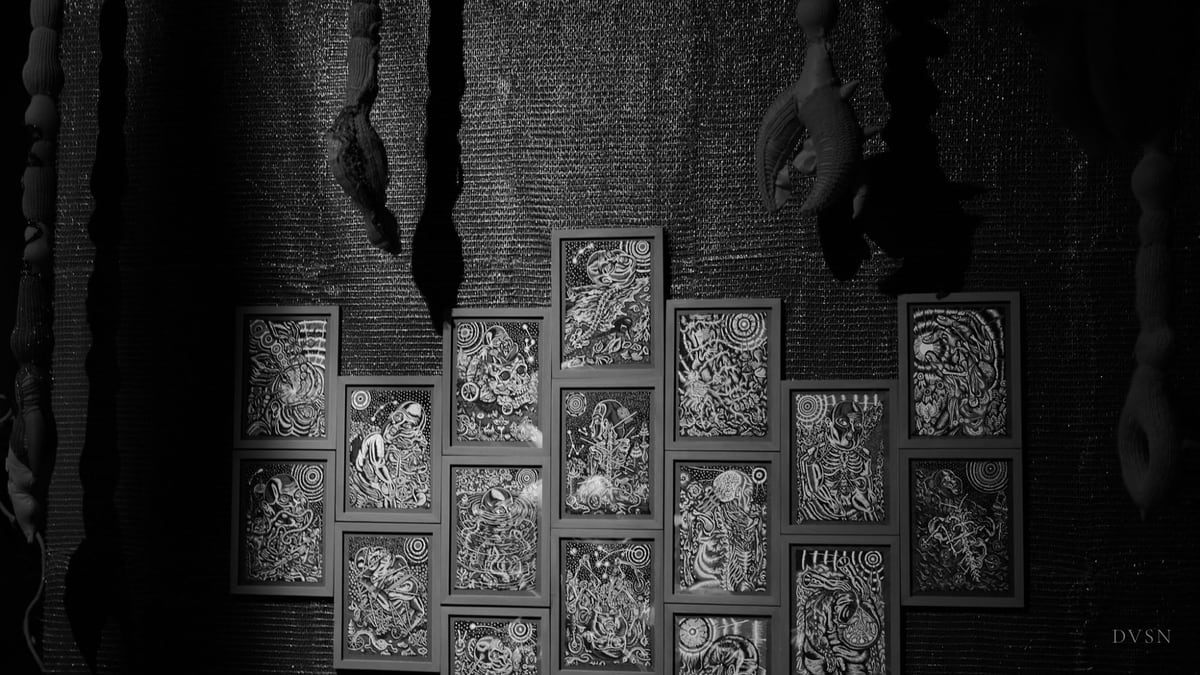 Un mur de dessins en noir et blanc encadrés, sous des formes textiles suspendues, dans la pénombre.