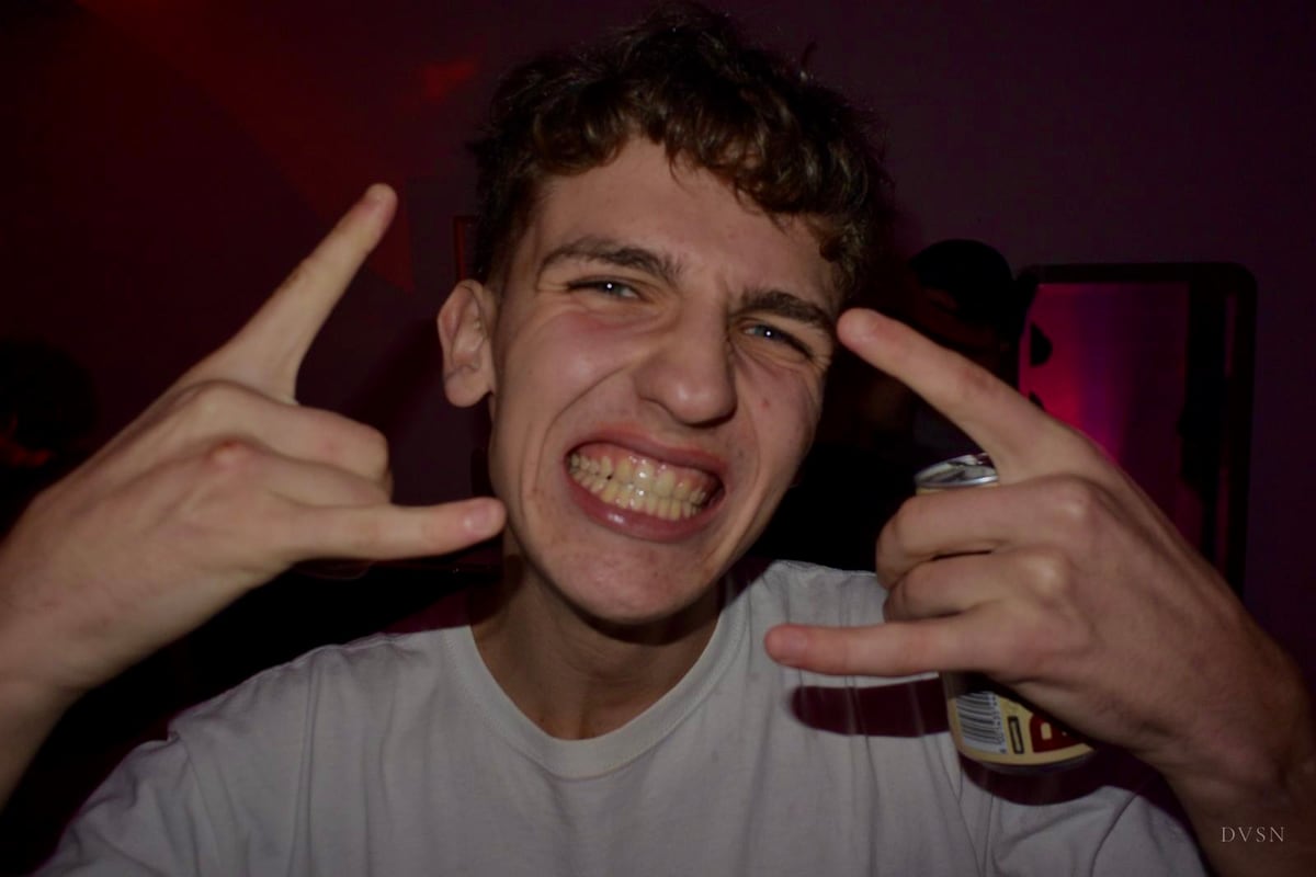 Un jeune homme grimace un sourire sous le flash, mains levées dans un geste joueur, une canette à la main.
