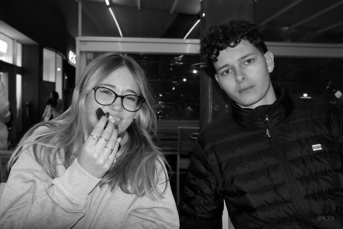 Deux amis en terrasse la nuit ; elle rit derrière sa main, il fixe calmement l'objectif.