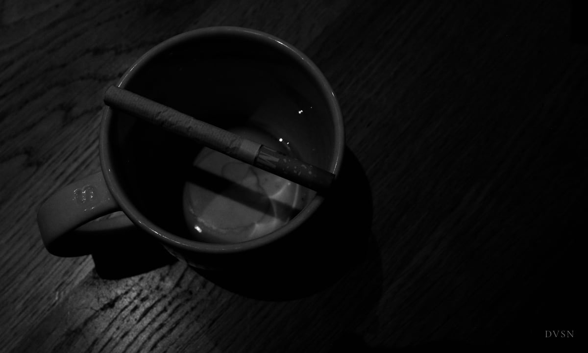 Une cigarette posée en travers d'une tasse vide sur une table en bois sombre, vue d'en haut.