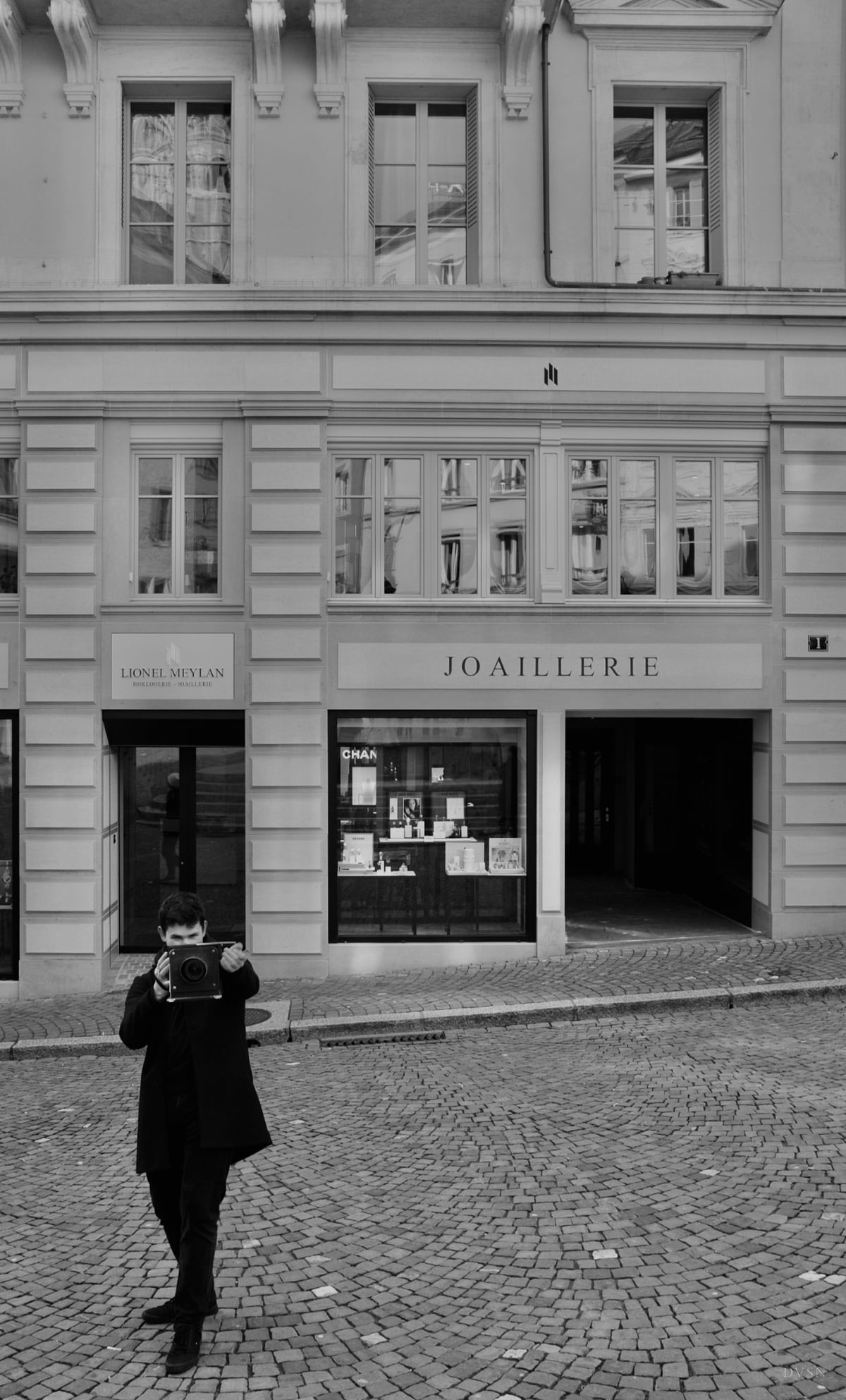 Un homme en manteau sombre pointe une chambre photographique vers le spectateur, devant une façade en pierre avec une joaillerie.